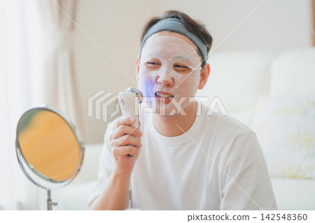 一位40歲的男子一邊使用臉部護理儀一邊敷面膜，臉上帶著笑容。男士美容。 142548360