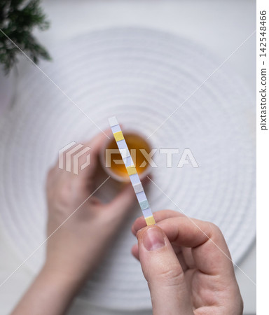 Person dipping a reagent test strip into a urine specimen container for medical analysis. Practical home diagnostic test checking health parameters and biomarkers. Person dipping a reagent test strip into a urine specimen container for medical analysis. Practical home diagnostic test checking health parameters and biomarkers. 142548466