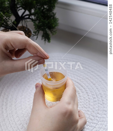 Person dipping a reagent test strip into a urine specimen container for medical analysis. Practical home diagnostic test checking health parameters and biomarkers. Person dipping a reagent test strip into a urine specimen container for medical analysis. Practical home diagnostic test checking health parameters and biomarkers. 142548488