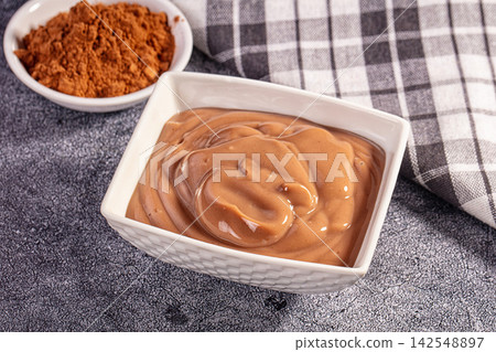 Smooth chocolate pudding in white bowl with cocoa powder and checkered cloth 142548897