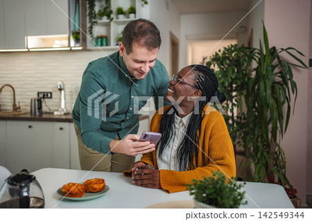 Interracial couple sharing cell phone content in kitchen Interracial couple sharing cell phone content in kitchen 142549344