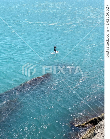 Lone paddleboarder crosses turquoise coastal water on bright sunny summer day. Lone paddleboarder crosses turquoise coastal water on bright sunny summer day. 142550627