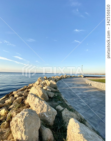 Rocky seaside promenade curves along calm water in warm evening sunlight. Rocky seaside promenade curves along calm water in warm evening sunlight. 142550810