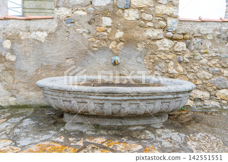 Ancient Stone Wall Fountain with Oval Basin and Brass Tap in Old European Village Street. Antique Open air bath tub with water Ancient Stone Wall Fountain with Oval Basin and Brass Tap in Old European Village Street. Antique Open air bath tub with water 142551551