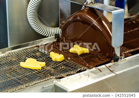 Marzipan candies on conveyer line being glazed with melted chocolate at sweet factory. Process of glazing. Chocolate tempering machine Marzipan candies on conveyer line being glazed with melted chocolate at sweet factory. Process of glazing. Chocolate tempering machine 142552045