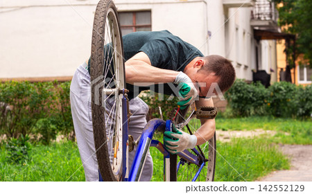 Man repairing bicycle upside down on sidewalk. Checking for hidden defects. DIY mechanical skills in action. Preparing two wheeler for upcoming long journey. Wheel alignment Man repairing bicycle upside down on sidewalk. Checking for hidden defects. DIY mechanical skills in action. Preparing two wheeler for upcoming long journey. Wheel alignment 142552129