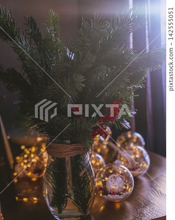 Christmas pine branches with red berries in glass vase, cozy festive lights background 142555051