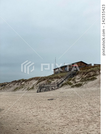 Sandy beach with wooden stairs leading up to a coastal house under overcast sky, minimal and calm seaside scene Sandy beach with wooden stairs leading up to a coastal house under overcast sky, minimal and calm seaside scene 142555423