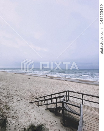 Empty sandy beach with wooden stairs leading to the sea under soft cloudy sky and calm waves 142555429