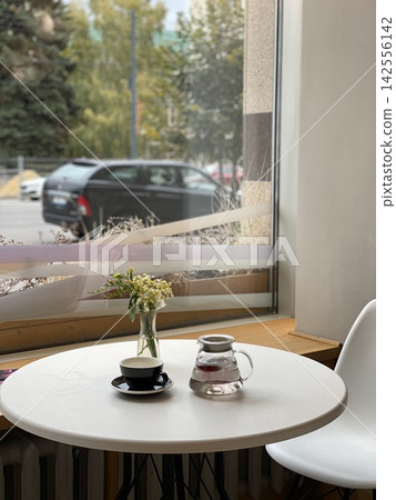 Minimalist cafe table by a large window with black coffee cup, glass teapot and small bouquet of yellow and white flowers. Minimalist cafe table by a large window with black coffee cup, glass teapot and small bouquet of yellow and white flowers. 142556142