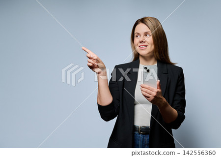 Adult businesswoman holding smartphone pointing finger away with copy space on grey background Adult businesswoman holding smartphone pointing finger away with copy space on grey background 142556360
