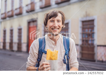 Man traveler holding a golden molote poblano traditional fried Mexican empanada outdoors on a street in Puebla Mexico with colonial city background paper wrapper local street food authentic travel. Man traveler holding a golden molote poblano traditional fried Mexican empanada outdoors on a street in Puebla Mexico with colonial city background paper wrapper local street food authentic travel. 142557032