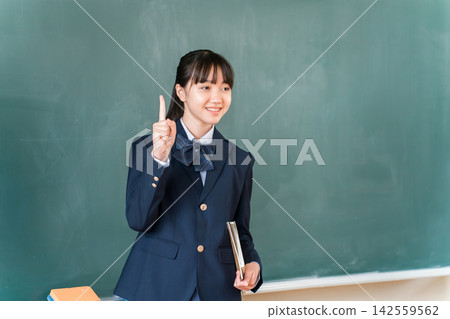 A high school girl smiles and points to an important element in front of the classroom blackboard. This image represents an educational setting where she gives advice on studying and exams. A high school girl smiles and points to an important element in front of the classroom blackboard. This image represents an educational setting where she gives advice on studying and exams. 142559562