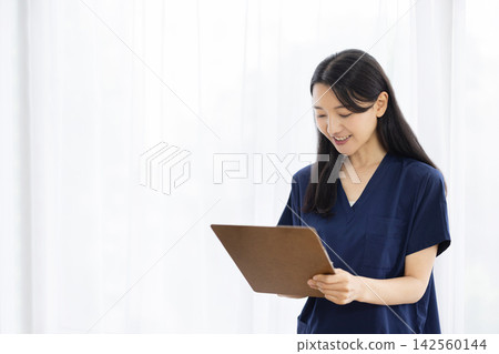 A young Japanese female doctor, wearing medical scrubs, takes notes in a binder by a bright window. A young Japanese female doctor, wearing medical scrubs, takes notes in a binder by a bright window. 142560144