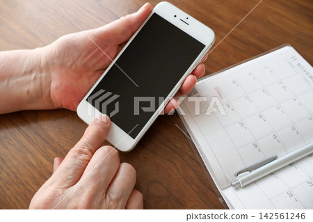 An elderly woman's hand holding a smartphone and a schedule book on the table. An elderly woman's hand holding a smartphone and a schedule book on the table. 142561246