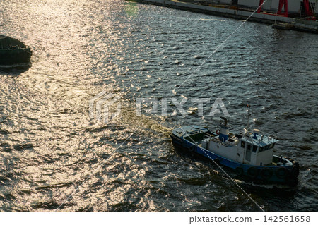 The silhouettes of tugboats and barges moving through the sparkling waters of Osaka South Port in the evening light. The silhouettes of tugboats and barges moving through the sparkling waters of Osaka South Port in the evening light. 142561658