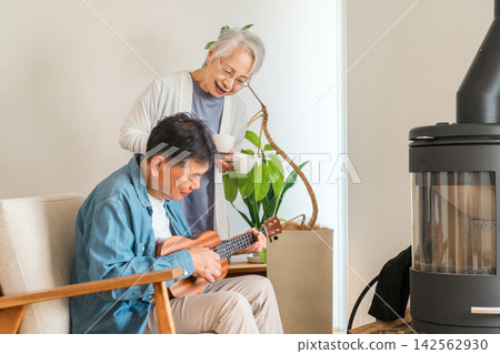 一對六、七十歲的老年夫婦在家中,圍坐在燃木爐旁,一邊彈著尤克里裡,一邊喝著咖啡。他們享受著自己的嗜好,過著充實而快樂的退休生活。 一對六、七十歲的老年夫婦在家中,圍坐在燃木爐旁,一邊彈著尤克里裡,一邊喝著咖啡。他們享受著自己的嗜好,過著充實而快樂的退休生活。 142562930