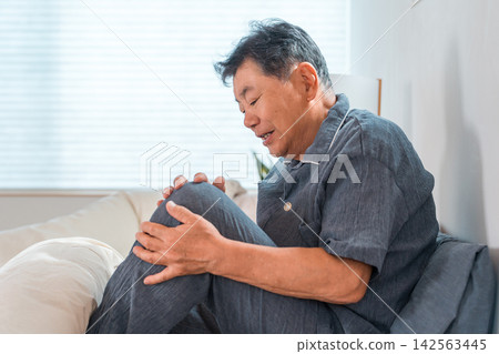 A senior man in his 60s or 70s sitting in bed, suffering from knee pain. This image represents joint pain and age-related knee problems. A senior man in his 60s or 70s sitting in bed, suffering from knee pain. This image represents joint pain and age-related knee problems. 142563445