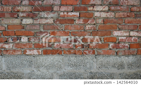 A section of an old wall combining two materials. The lower part is constructed of gray, porous-textured cinder blocks. Above lies uneven red brickwork featuring chipped bricks and gray mortar. A section of an old wall combining two materials. The lower part is constructed of gray, porous-textured cinder blocks. Above lies uneven red brickwork featuring chipped bricks and gray mortar. 142564010