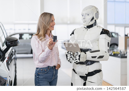 A woman holds a car key while interacting with a humanoid robot holding a tablet in a modern car showroom A woman holds a car key while interacting with a humanoid robot holding a tablet in a modern car showroom 142564587