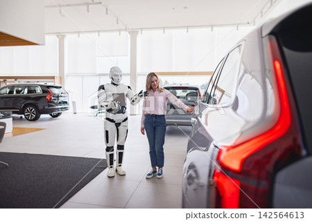 A humanoid robot holds a tablet and guides a woman as she opens the door of a new car in a dealership showroom A humanoid robot holds a tablet and guides a woman as she opens the door of a new car in a dealership showroom 142564613