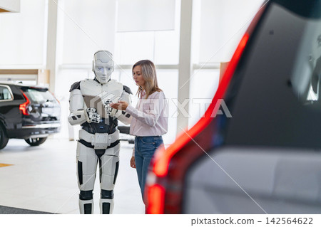 A humanoid robot interacts with a woman in a car showroom, holding a tablet and discussing vehicle options 142564622