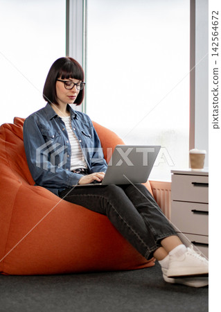 A young woman with glasses and a bob haircut sits comfortably on an orange bean bag chair, typing on her laptop with a coffee cup nearby 142564672