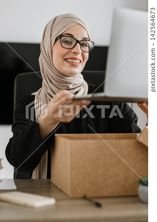 A smiling woman wearing a hijab and glasses is unboxing a laptop from a cardboard box at her desk A smiling woman wearing a hijab and glasses is unboxing a laptop from a cardboard box at her desk 142564673