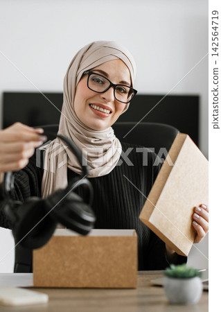 A smiling woman wearing a hijab and glasses opens a package containing headphones, showcasing a modern unboxing experience A smiling woman wearing a hijab and glasses opens a package containing headphones, showcasing a modern unboxing experience 142564719
