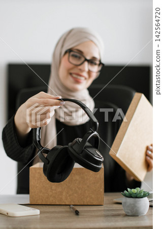 A smiling woman wearing a hijab and glasses is unboxing a new pair of black headphones from a cardboard package A smiling woman wearing a hijab and glasses is unboxing a new pair of black headphones from a cardboard package 142564720