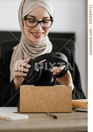 A smiling woman wearing a hijab and glasses unboxes a pair of black headphones from a cardboard package A smiling woman wearing a hijab and glasses unboxes a pair of black headphones from a cardboard package 142564721