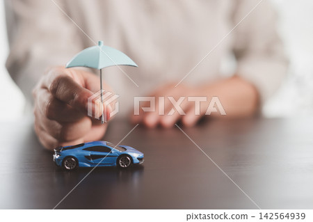 Person sheltering a blue model car with a small umbrella, symbolizing auto insurance, vehicle protection, financial security, risk management, accident coverage, and safe ownership. Person sheltering a blue model car with a small umbrella, symbolizing auto insurance, vehicle protection, financial security, risk management, accident coverage, and safe ownership. 142564939