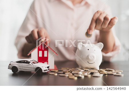 Person dropping a coin into a white piggy bank beside a model house, car, and scattered coins, symbolizing savings, budgeting, home and vehicle goals, financial planning, and future security. Person dropping a coin into a white piggy bank beside a model house, car, and scattered coins, symbolizing savings, budgeting, home and vehicle goals, financial planning, and future security. 142564940
