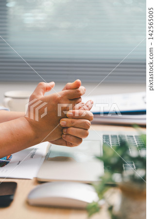Office worker clasping hands above laptop and financial report at desk, showing a brief work break, hand stretching, stress relief, workplace wellness, ergonomics and healthy productivity routine. Office worker clasping hands above laptop and financial report at desk, showing a brief work break, hand stretching, stress relief, workplace wellness, ergonomics and healthy productivity routine. 142565005