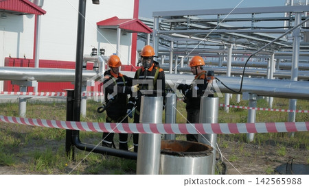Three male workers in orange helmets check metal pipes at industrial plant, with one worker using gas mask. Suspenseful refinery maintenance takes place behind safety tape during safety inspection. Three male workers in orange helmets check metal pipes at industrial plant, with one worker using gas mask. Suspenseful refinery maintenance takes place behind safety tape during safety inspection. 142565988