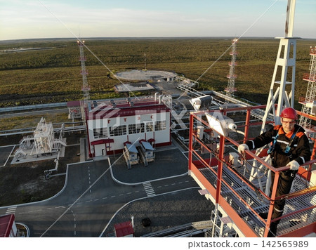 Industrial worker performs routine inspection on elevated platform, wearing red helmet, reflective coveralls, gloves, and harness. Processing equipment, pipework, towers, and open tundra extend Industrial worker performs routine inspection on elevated platform, wearing red helmet, reflective coveralls, gloves, and harness. Processing equipment, pipework, towers, and open tundra extend 142565989