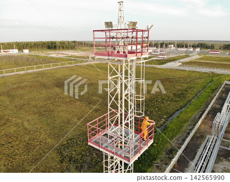 Industrial worker in orange safety gear stands on elevated tower platform, holding red railing beside mounted floodlights. Hard hat, guardrails, ladders, pipes, and distant facility emphasize secure Industrial worker in orange safety gear stands on elevated tower platform, holding red railing beside mounted floodlights. Hard hat, guardrails, ladders, pipes, and distant facility emphasize secure 142565990