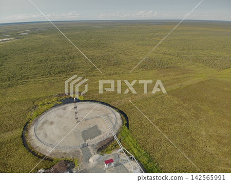 Remote industrial facility sits on circular concrete pad with fence, pipes, and small service building. Surrounding marshy grassland spreads across wide horizon, giving isolated utility site context. Remote industrial facility sits on circular concrete pad with fence, pipes, and small service building. Surrounding marshy grassland spreads across wide horizon, giving isolated utility site context. 142565991