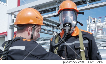 Two male industrial workers in orange helmets wear respirators at plant, one facing camera with focused expression. Safety gear and steel framework suggest industrial style at refinery or factory Two male industrial workers in orange helmets wear respirators at plant, one facing camera with focused expression. Safety gear and steel framework suggest industrial style at refinery or factory 142565992