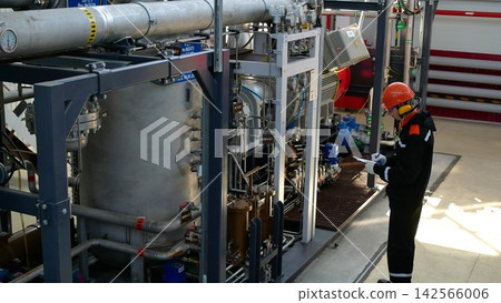 Industrial technician writes on clipboard beside large pipe assembly, focused in protective helmet and gloves. Industrial technician stands in futuristic plant, checking gauges and valves on metal Industrial technician writes on clipboard beside large pipe assembly, focused in protective helmet and gloves. Industrial technician stands in futuristic plant, checking gauges and valves on metal 142566006