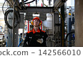 Factory worker reviews clipboard beneath complex machinery, focused young adult man in safety helmet, eye protection, earmuffs, and gloves works inside industrial plant with stainless steel piping. 142566010