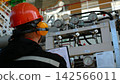Back view of maintenance worker in hard hat reviews gauge readings at industrial panel, focused and attentive. Worker stands before pipes, dials, and clipped paperwork, inspecting meters and valves. 142566011