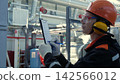 Technician in safety gear records notes on clipboard inside industrial facility, with concentrated expression while checking equipment, pipes, and metal ducts around work area during inspection. 142566012