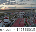 Fuel processing facility contains large white storage tanks, red roofed industrial buildings, extensive metal piping, and tall lattice towers. Flat marshland and sunset horizon surround fenced plant 142566015
