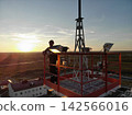 Maintenance worker stands inside elevated red metal tower on rooftop. Bright setting sun lights antenna structure, cables, and lamps while flat fields stretch across distant horizon. 142566016