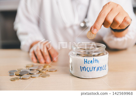 Healthcare professional placing a coin into a jar labeled Health Insurance, symbolizing medical savings, insurance premiums, healthcare budgeting, financial protection and future medical expenses. Healthcare professional placing a coin into a jar labeled Health Insurance, symbolizing medical savings, insurance premiums, healthcare budgeting, financial protection and future medical expenses. 142569968
