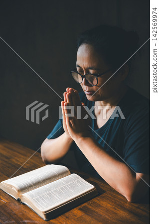 Adult person with glasses praying with closed eyes and palms together in a dark room, expressing faith, devotion, worship, spirituality, hope, peace, reflection, gratitude, and quiet meditation. Adult person with glasses praying with closed eyes and palms together in a dark room, expressing faith, devotion, worship, spirituality, hope, peace, reflection, gratitude, and quiet meditation. 142569974