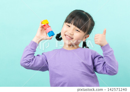 A cute young student holding up a stack of colorful building blocks while giving a confident thumbs up with her other hand 142571240