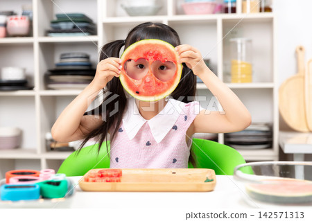 Playful young Asian girl covering one eye with watermelon slice while holding heart shaped toy in other hand Playful young Asian girl covering one eye with watermelon slice while holding heart shaped toy in other hand 142571313