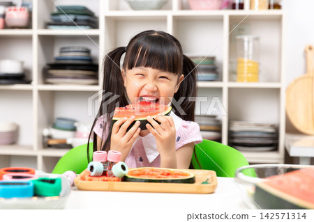 Cheeky young Asian girl while holding watermelon slice at kitchen table with bright smile Cheeky young Asian girl while holding watermelon slice at kitchen table with bright smile 142571314